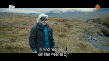 Britta Marakatt bij een stroompje waar ze al een tijd niet was geweest. Close-up van een witharige vrouw met bril en een donkerblauwe haarband om haar voorhoofd en oren. Ze draagt een lichtblauwe (dichte) jas met daaroverheen een donkerblauwe (open) jas. Rond haar nek, half onder de lichtblauwe jas draagt ze een veelkleurige das. De vrouw staat op een toendra met droog gras, op de achtergrond zijn bergen en mist te zien. De ondertiteling zegt: "Ik vind het heel fijn om hier weer te zijn."